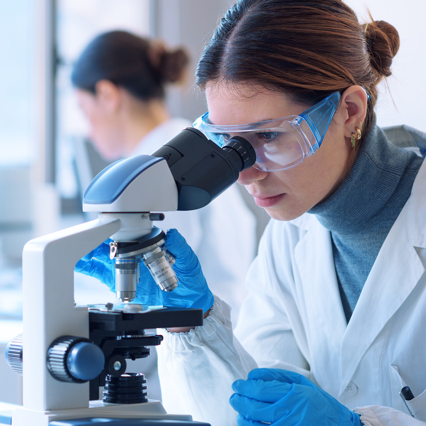 Ricercatrice con occhiali protettivi osserva al microscopio in laboratorio · Euroclone